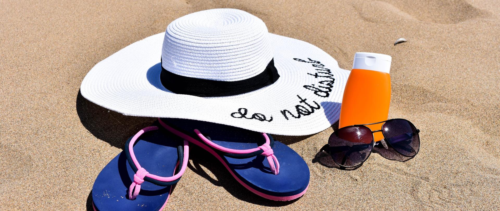 Na piasku na plaży ułożony kapelusz, krem, okulary i klapki.