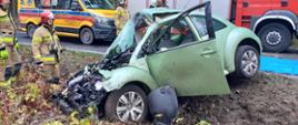 Rozbity przód samochodu osobowego w przydrożnym rowie. W tle wóz strażacki, karetka pogotowia i stojący obok strażacy