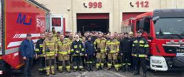 Zdjęcie grupowe uczestników szkolenia pomiędzy dwoma samochodami pożarniczymi na tle remizy strażackiej