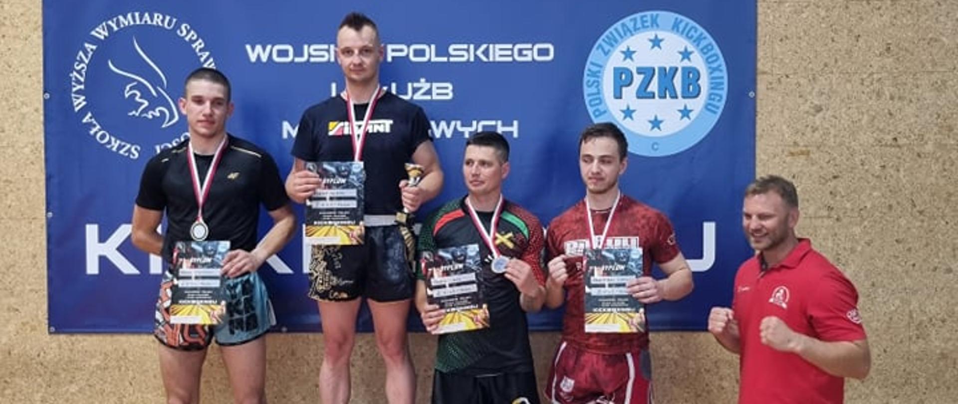 Adrian Durma stojący na pierwszym miejscu podium, obok pozostali zawodnicy. Za ich plecami baner z napisem Puchar Polski Wojska Polskiego i Służb Mundurowych w kickboxingu.