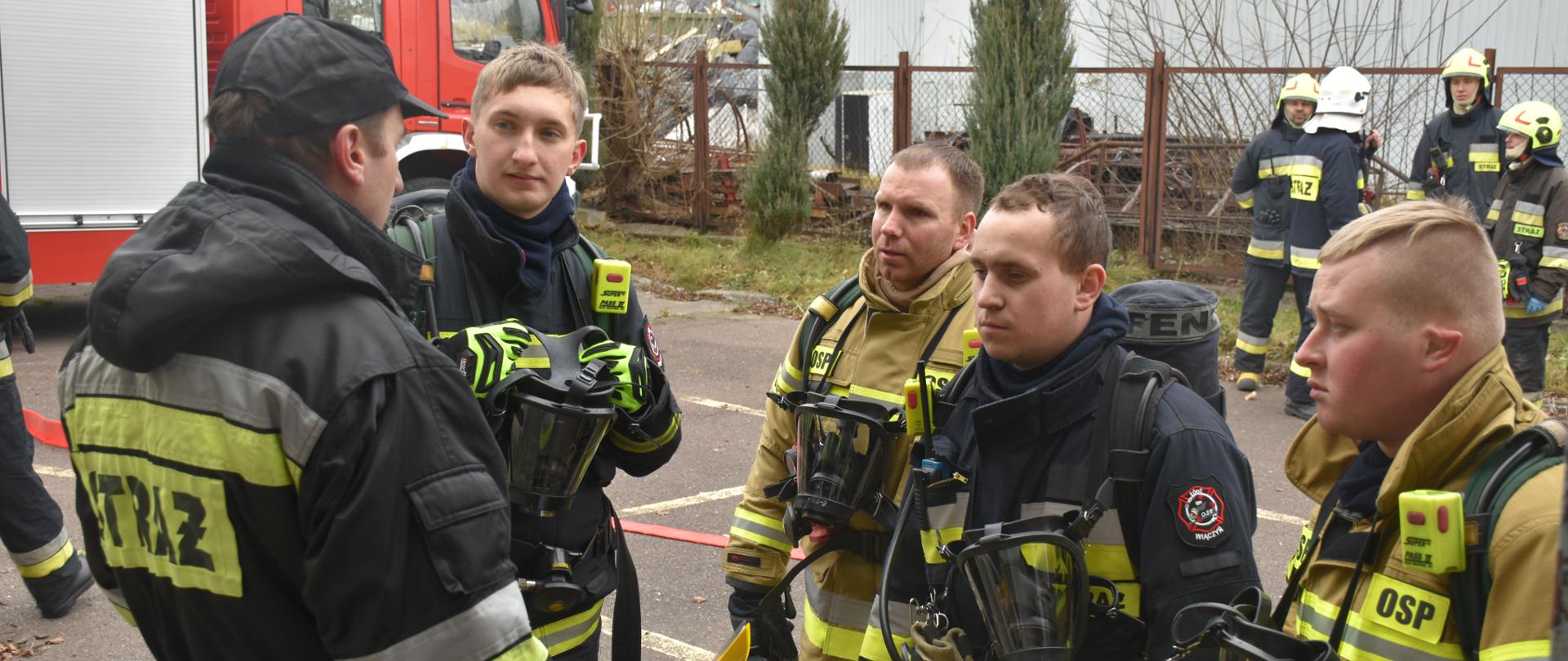 Czterech strażaków w ubraniach specjalnych z aparatami powietrznymi na plecach oraz maskami ochrony dróg oddechowych zawieszonymi na szyjach. Przed nimi strażak podczas odprawy przed egzaminem ubrany w strój dowódczo-sztabowy. W tle czerwony samochód pożarniczy oraz czterech strażaków w ubraniach specjalnych i hełmach na głowach.