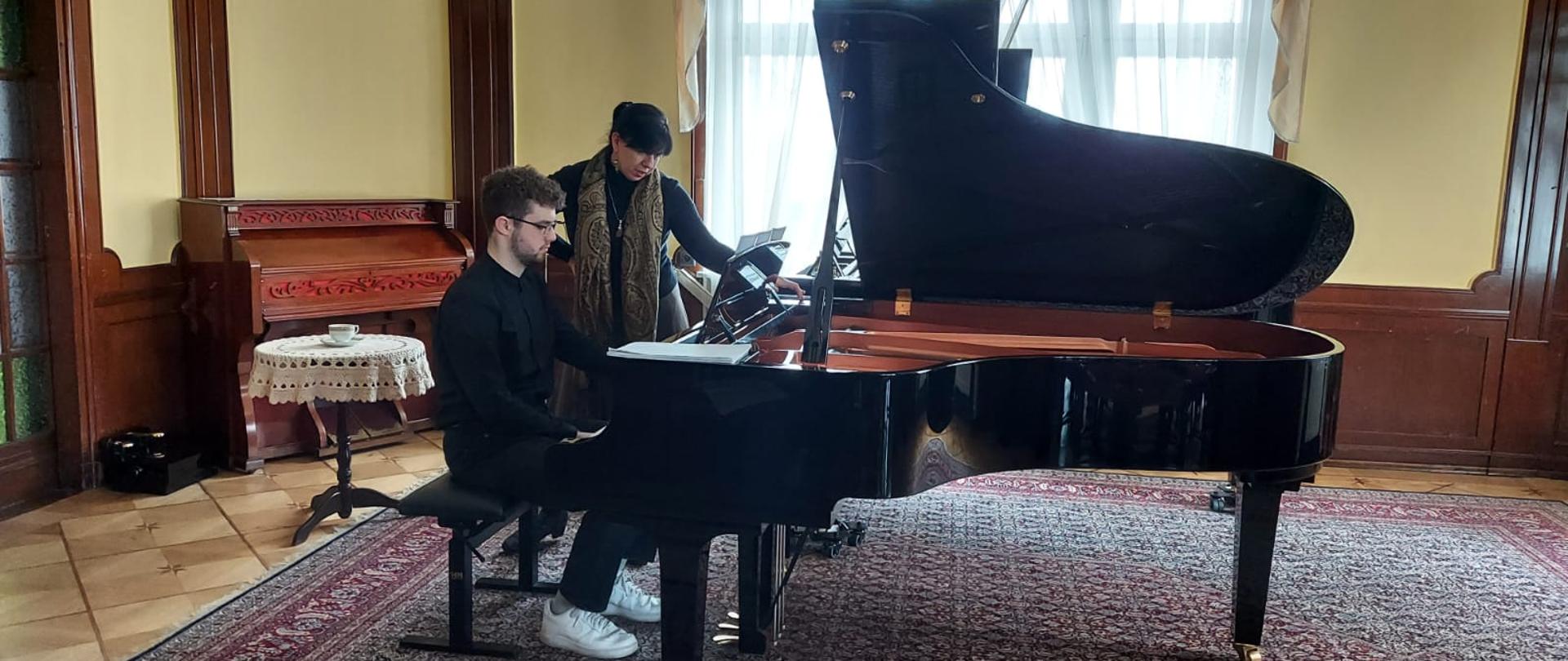 Zdjęcie kolorowe. Uczeń w czarnym stroju przy fortepianie z prof. J. Marcinkowską gra. 