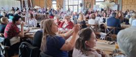 Zdjęcie przedstawia wnętrze dużej, elegancko zaaranżowanej sali, w której odbywa się spotkanie o charakterze konferencyjno integracyjnym. W pomieszczeniu ustawiono liczne drewniane stoły i krzesła, przy których zasiadają uczestnicy wydarzenia. Sale wypełnia duża liczba osób, rozmieszczonych równomiernie w przestrzeni, co wskazuje na wysoką frekwencję.