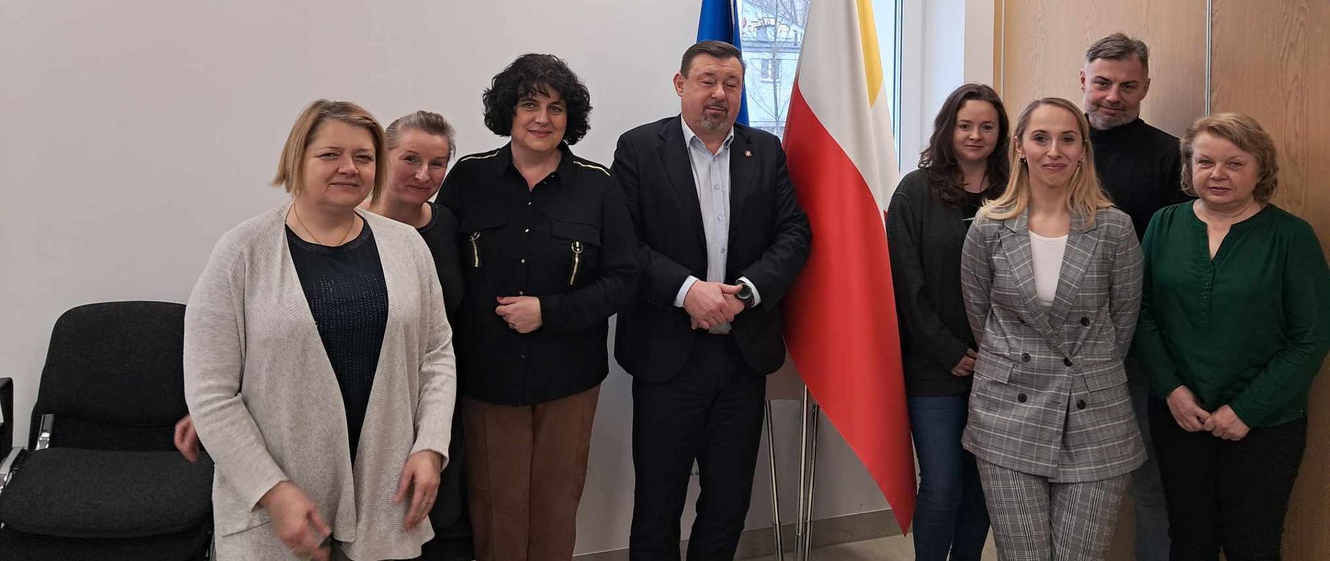 Dziewięć osób stoi w pokoju. Za nimi znajdują się trzy flagi