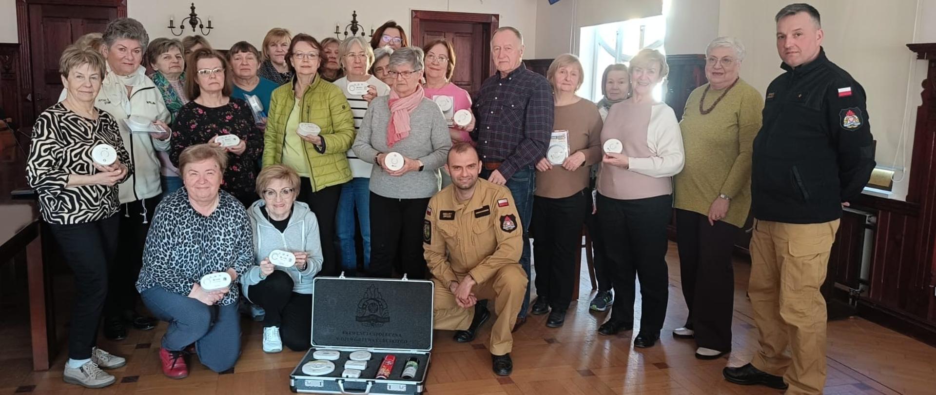 Zdjęcie przedstawia grupę uczestników spotkania wraz ze strażakami. Uczestnicy spotkania trzymają w dłoniach czujniki.