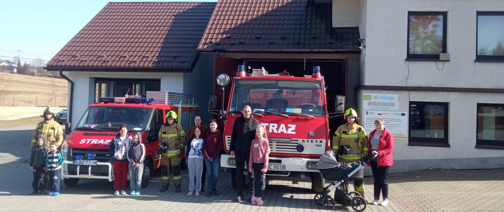 Zdjęcie przedstawia w centralnej części grupę ludzi - strażaków oraz obywateli Ukrainy. Ludzie stoją przy czerwonych samochodach pożarniczych. W dalszym planie widać budynek remizy OSP Wiśnicz Mały.