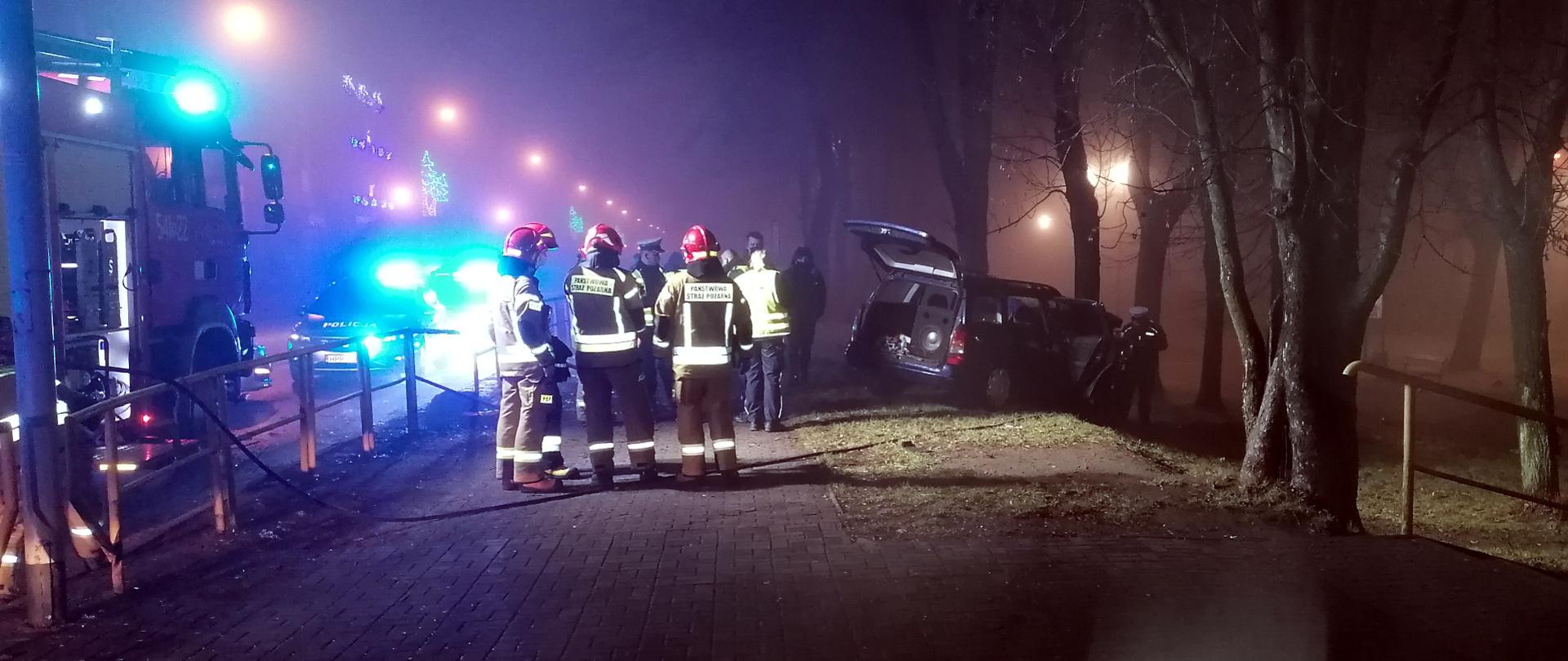 Zdjęcie przedstawia z lewej strony częściowo widoczny średni samochód ratowniczo - gaśniczy, po środku grupę strażaków podczas działań, a z prawej strony granatowy samochód kombi z otwartą tylną klapą, wbity w drzewo. W tle drzewa i światła ostrzegawcze straży pożarnej i policji.