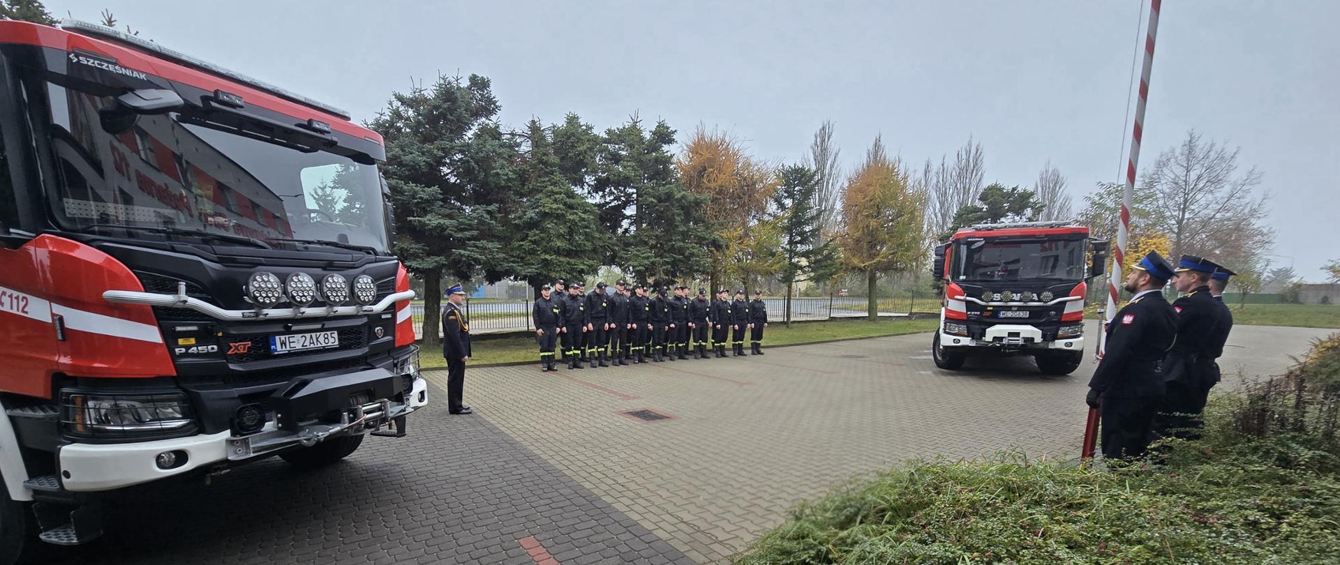 W centralnej części zdjęcia strażacy w ubraniach koszarowych w dwuszeregu, po ich lewej i prawe stronie wozy strażackie. Uroczystość odbywa się przed budynkiem komendy 