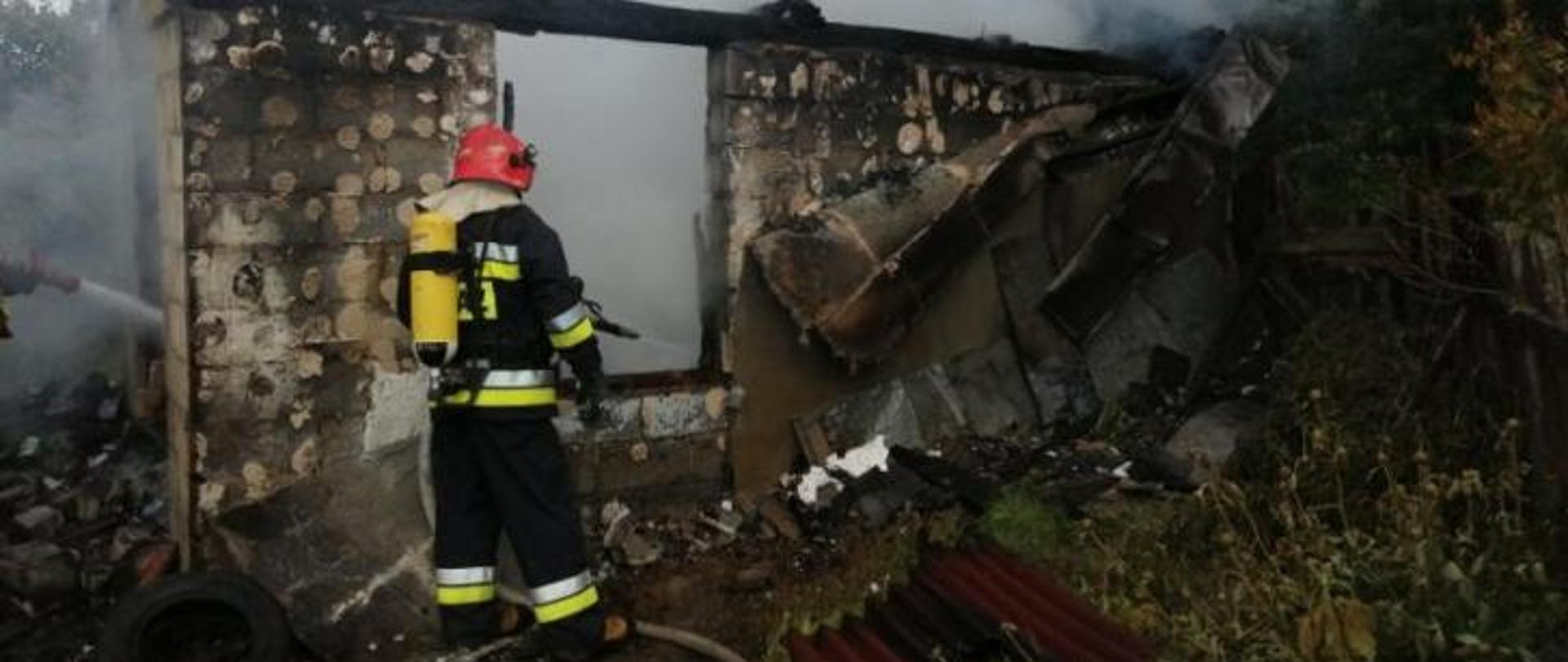 Zdjęcie przedstawia strażaka w ubraniu bojowym dogaszającego pogorzelisko pożaru budynku.