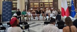 Uczestnicy Narodowego Czytania siedzą w półkolu w Bibliotece Narodowej Malty, na tle księgozbiorów. Przed nimi siedzą osoby z widowni.
