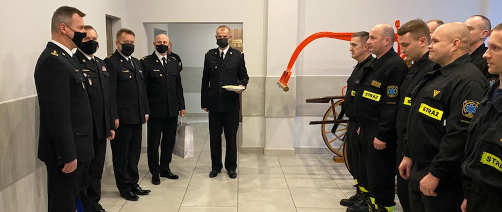 Zdjęcie przedstawia 10 strażaków podczas spotkania w sali w jednostce ratowniczo-gaśniczej PSP. Po lewej stronie stoi pięciu strażaków (kadra kierownicza PSP. Po lewej stronie stoi pięciu strażaków w szeregu. 