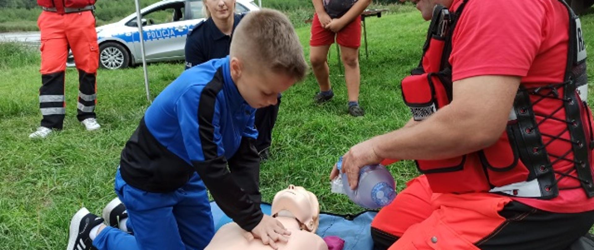 na pierwszym planie jest chłopiec, który ćwiczy resuscytację na fantomie oraz ratownik medyczny. W tle samochód pożarniczy i radiowóz oraz policjantka i ratownik medyczny. 