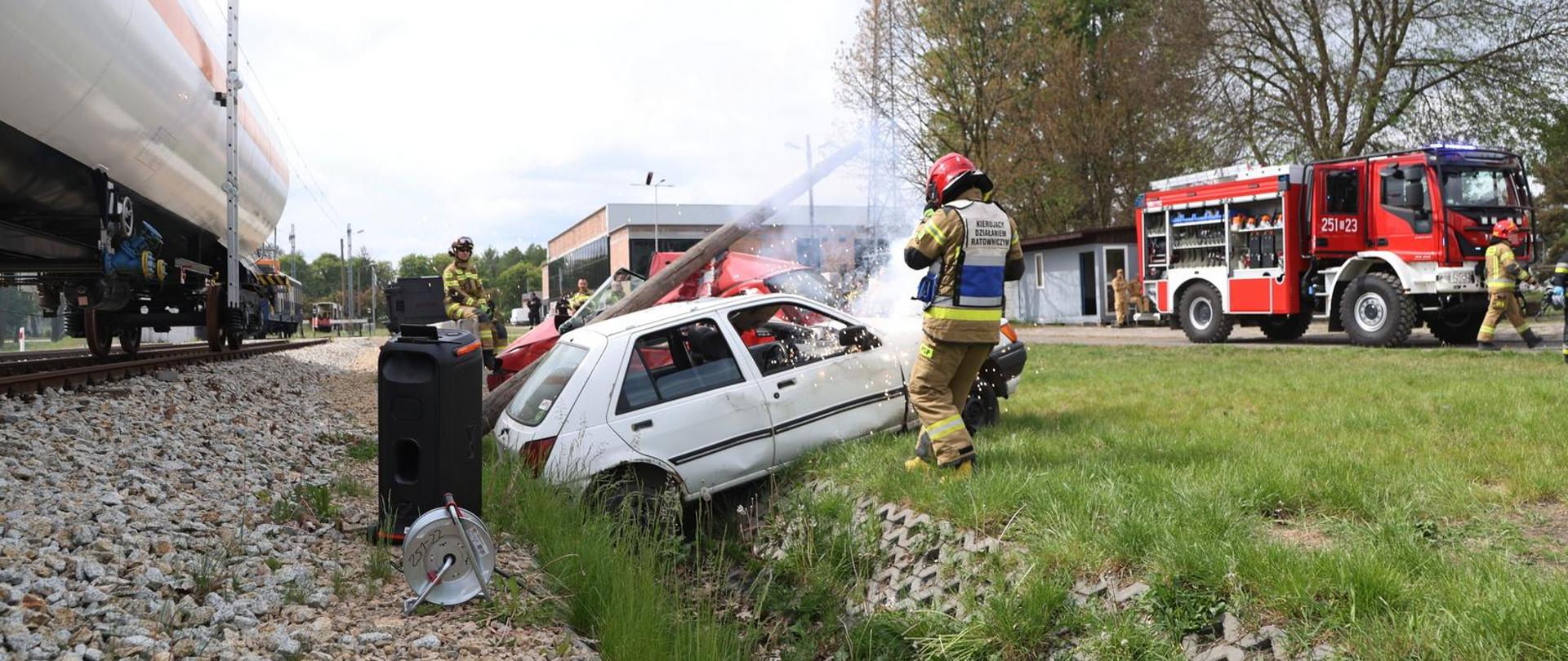 Poligon Centralnej Szkoły PSP, wraki samochodów osobowych ułożone w sposób symulujący zdarzenie drogowe, w głębi po prawej bojowy samochód pożarniczy