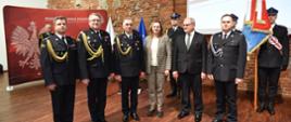 trzech strażaków, kobieta, mężczyzna i strażak ochotnik pozują do zdjęcia w sali konferencyjnej, w tle ścianka wspinaczkowa i poczet sztandarowy