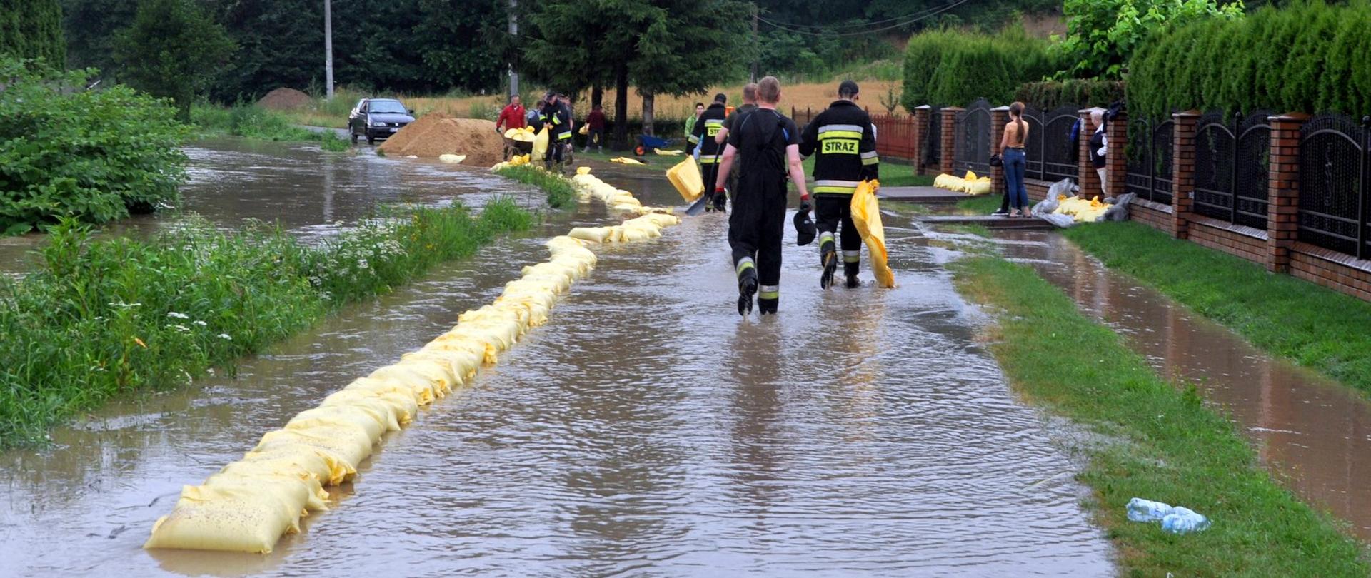 Na zdjęciu strażacy, odwróceniu tyłem niosą worki wypełnione piaskiem do zabezpieczenia posesji przed zalaniem. Po lewej stronie widać ułożone worki z piaskiem.