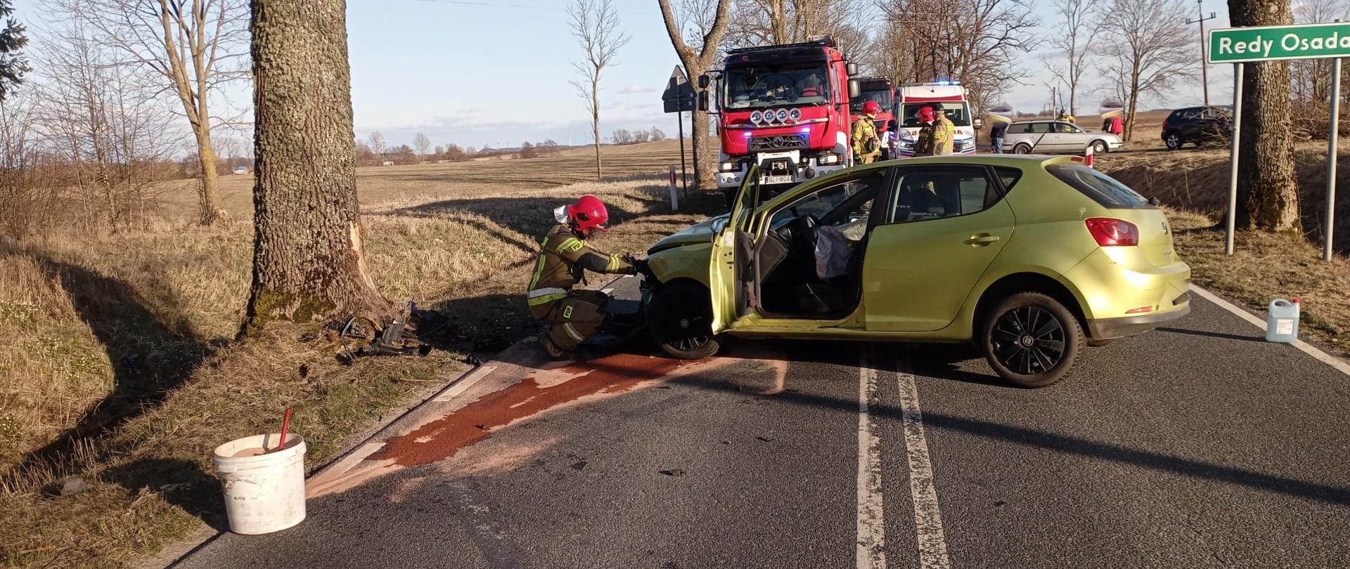 Zdjęcie przedstawia uszkodzony pojazd po czołowym uderzeniu w przydrożne drzewo oraz strażaka PSP, który próbuje otworzyć maskę do komory silnika. 