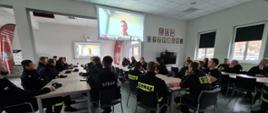 Na zdjęciu strażacy przy stole w tle widoczna jest wyświetlona na ekranie Video Konferencja