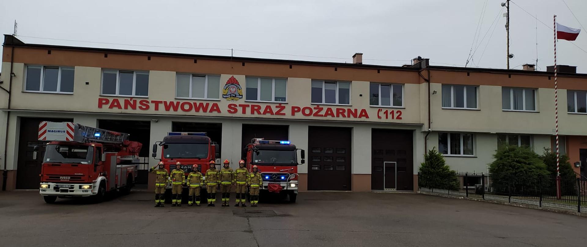 Strażacy oraz samochody ratowniczo-gaśnicze stoją przed garażami z włączonymi sygnałami świetlno-dźwiękowymi.
