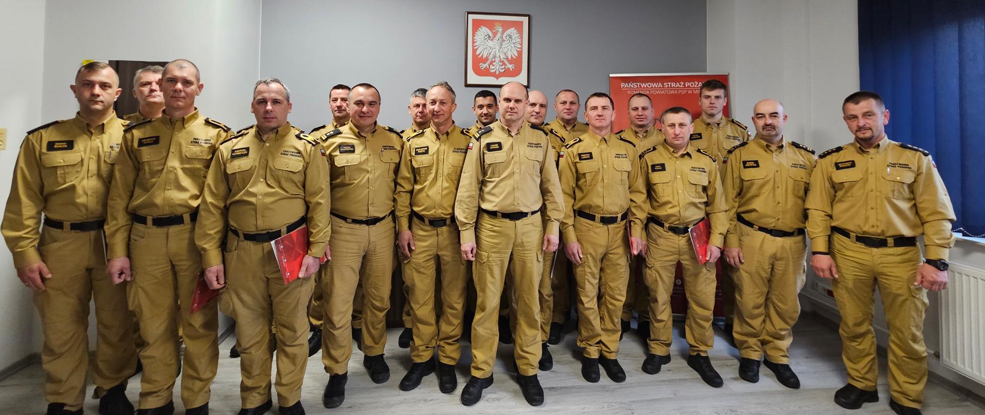 Na zdjęciu znajduje się grupa kilkunastu mężczyzn, funkcjonariuszy Państwowej Straży Pożarnej stojących w rzędzie wewnątrz pomieszczenia. Wszyscy mężczyźni ubrani są w jednolite, beżowe mundury z ciemnymi elementami. Na ich mundurach widoczne są różne naszywki i oznaczenia stopni. Niektórzy z mężczyzn trzymają w rękach czerwone teczki lub dokumenty.
Mężczyźni stoją na tle jasnych ścian. Na ścianie za nimi wisi oprawione godło Polski - biały orzeł na czerwonym tle. Po prawej stronie widoczny jest fragment czerwonego banera z białym napisem "PAŃSTWOWA STRAŻ POŻARNA".
Zdjęcie wykonane podczas uroczystej zbiórki z okazji wręczenia decyzji odnośnie awansów na wyższe stanowiska służbowe oraz powitania nowego funkcjonariusza.