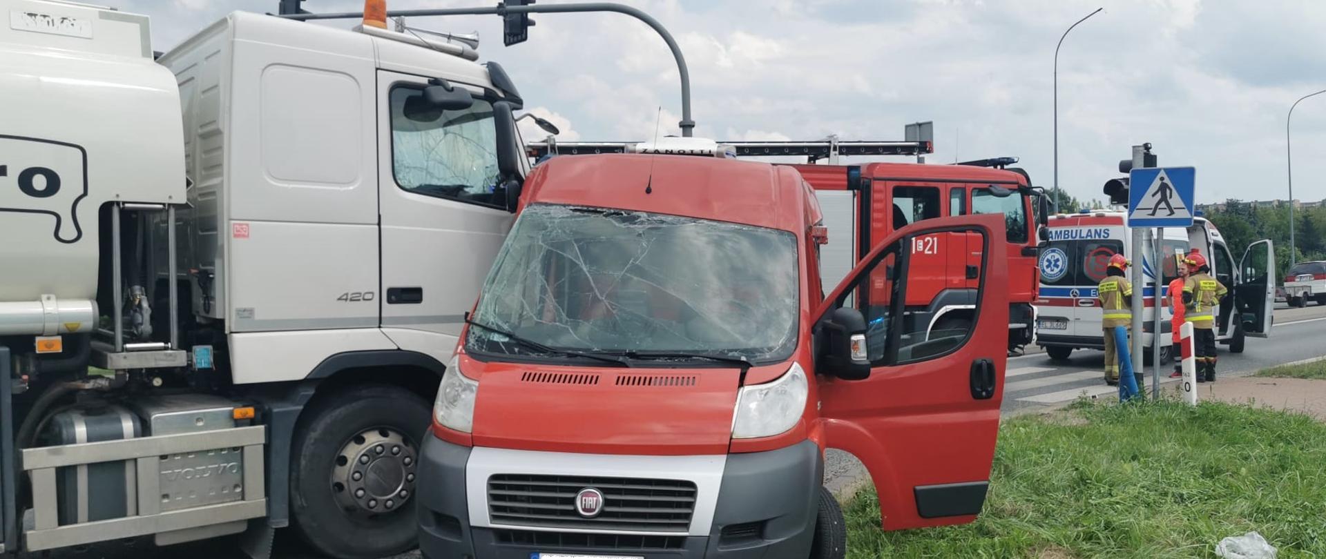 Zdjęcie przedstawia białą cysternę, która wjechała w bok czerwonego busa, na skrzyżowaniu ze światłami. Z tylu wiadać strażaków oraz karetkę pogotowia.