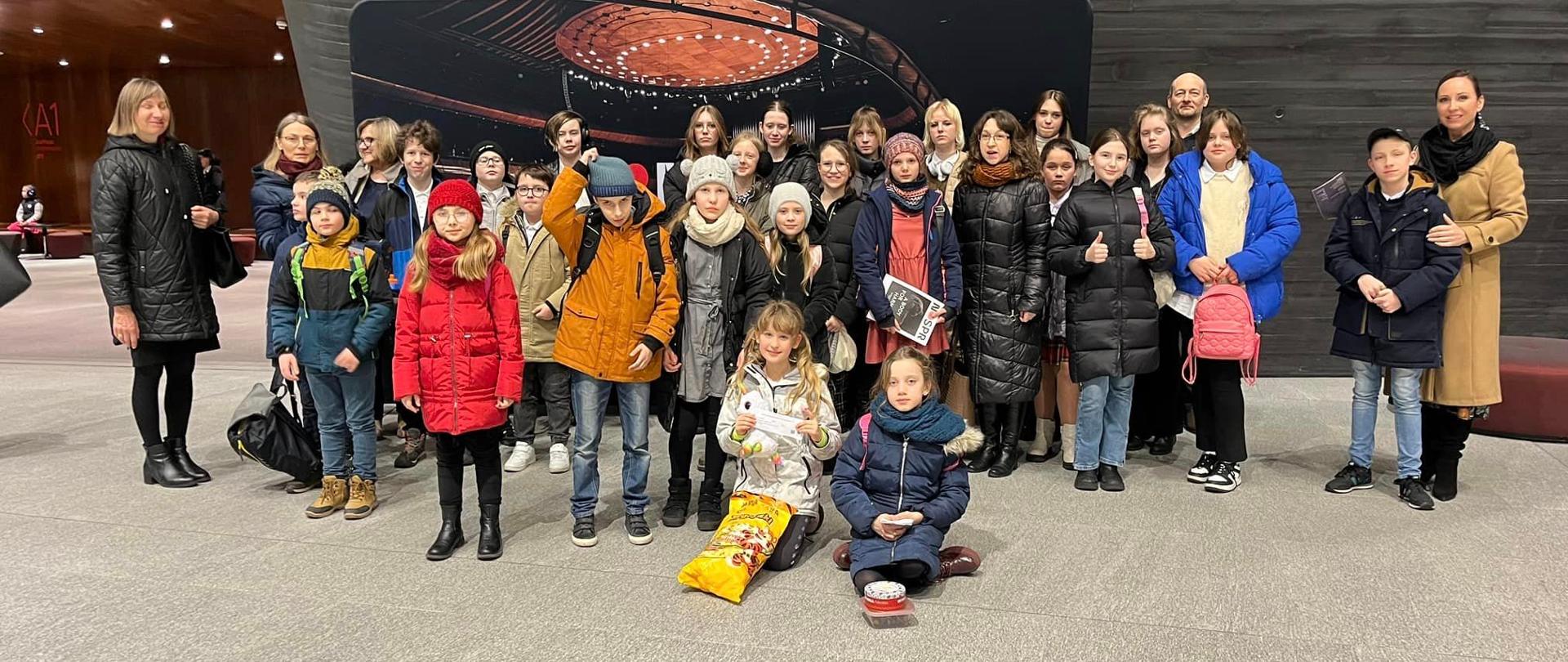 Zdjęcie grupy uczniów w NOSPR w Katowicach. Przedstawia uczniów i nauczycieli na tle ściany koloru szarego. Na pierwszym planie widać podłogę koloru beżowego.