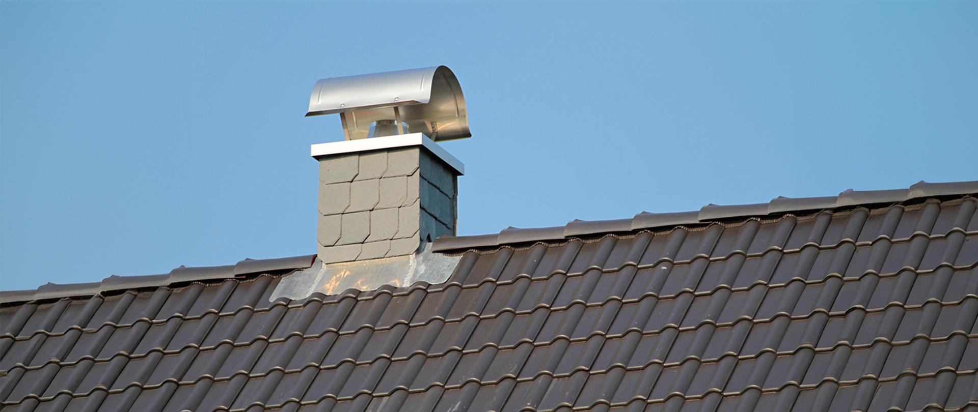 Na zdjęciu widać dach budynku pokryty ciemną dachówką ceramiczną. Na jego powierzchni znajduje się murowany komin wykończony jasnoszarą obudową oraz wyposażony w nowoczesną, metalową nasadę kominową w kształcie półokrągłej osłony. W tle widoczne jest czyste, błękitne niebo.