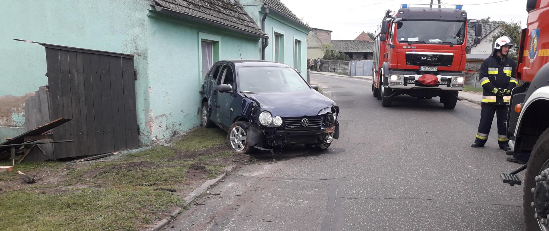 Na pierwszym planie rozbity samochód osobowy który chwilę temu wjechał w dom oraz ogrodzenie. Po prawej stronie dwa wozy strażackie a także strażak który zabezpiecza miejsce zdarzenia.