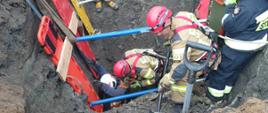 Na zdjęciu na widać wykop, na samym dole ledwo widoczna głowa poszkodowanego, strażacy zabezpieczają wykop i próbują odkopać zasypanego. Widać stabilizatory, linki i deski. 