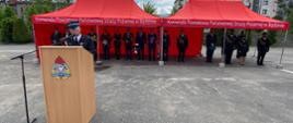 Zdjęcie przedstawia Komendanta Powiatowego Państwowej Straży Pożarnej w Będzinie mł. bryg. Arkadiusza Spera ubranego w mundur wyjściowy stojącego za mównicą podczas przemówienia. W tle pod czerwonym namiotem stoją wszyscy zaproszeni goście i cześć pracowników Komendy. Zdjęcie wykonano na placu Komendy.