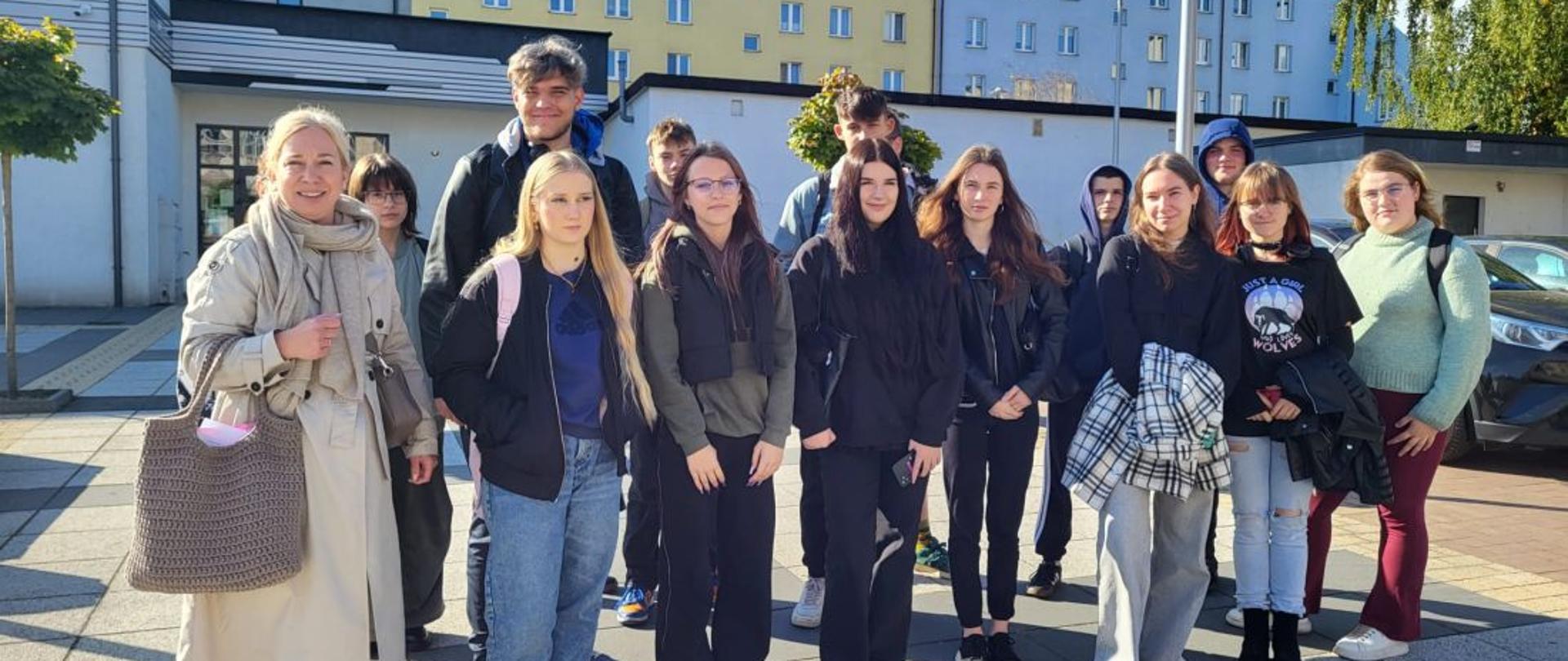 Grupa młodzieży z opiekunką pozują stojąc na placu, za nimi budynki