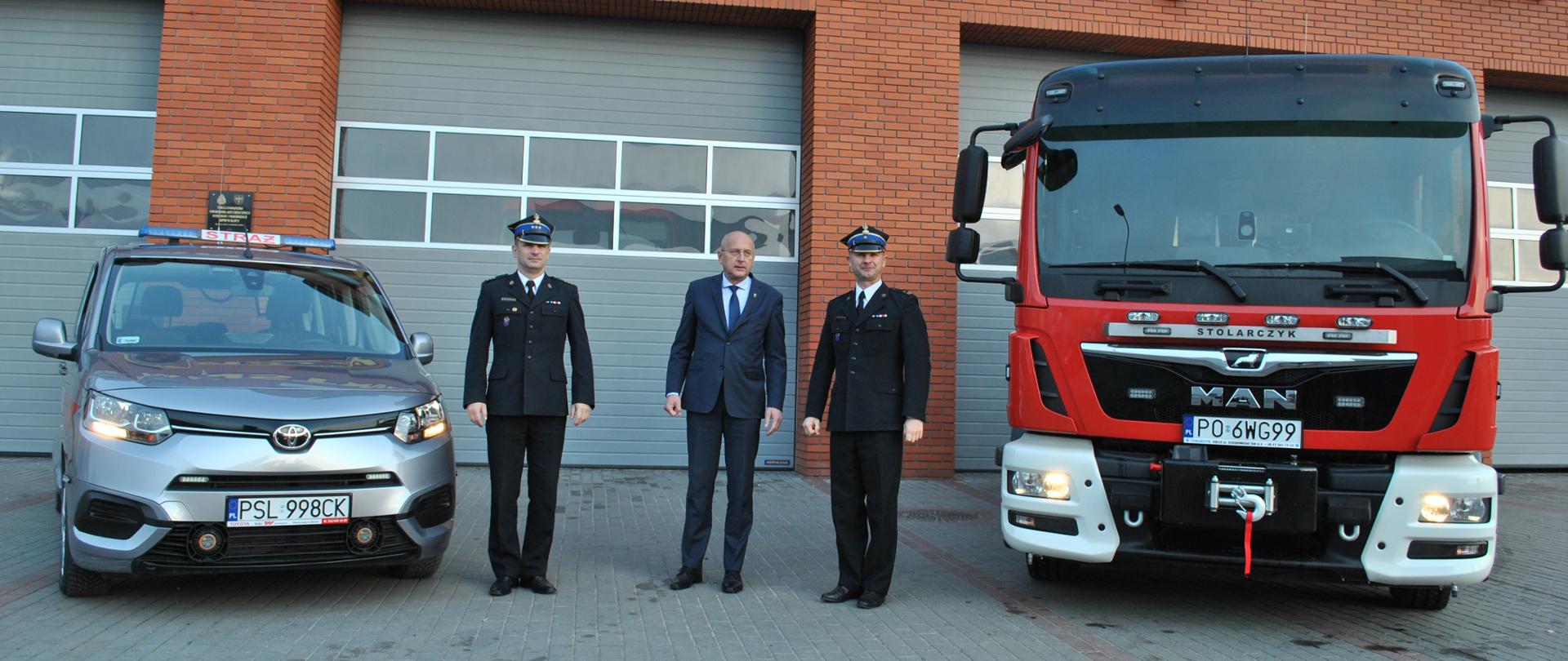 Dwóch funkcjonariuszy stoi przed budynkiem Państwowej Straży Pożarnej z jednej strony czerwony samochód marki MAN z drugiej strony srebrny samochód osobowy Toyota pomiędzy strażakami osoba w granatowym garniturze. Za nim bramy garażowe oraz budynek z cegły klinkier