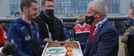 Na zdjęciu wojewoda opolski z jednym z zawodników ZAKSY. Wojewoda wręcza sportowcowi tort w pudełku z napisem zaksa. W tle prezes ZASKY Sebastian Świderski, patrzący w dal. 