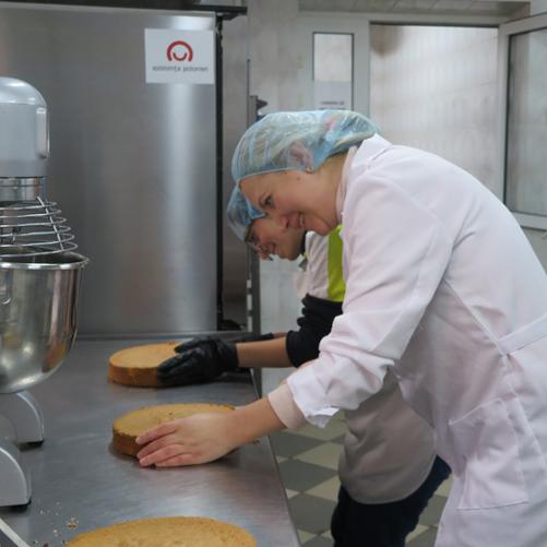 Dwie kobiety przygotowują ciasta w przemysłowej kuchni, w tle widać naklejkę z logo Polska pomoc