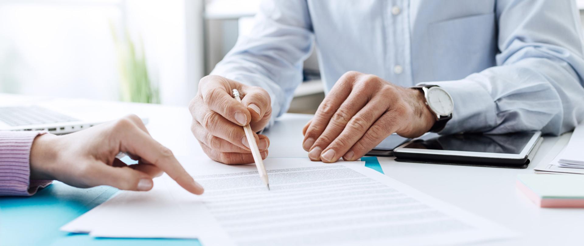 Ręce osób przy stole, jedna wskazuje ołówkiem na miejsce w dokumencie.