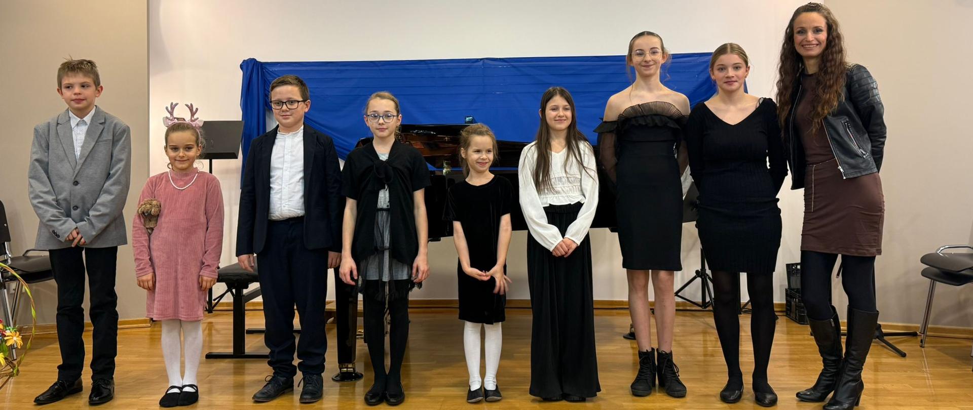 Dziesięć osób stoi w jednym rzędzie na podłodze z jasnego drewna w sali kameralnej. Wśród nich jest kilkoro dzieci i młodzież w wieku szkolnym oraz dwie dorosłe kobiety. W tle znajduje się fortepian przykryty niebieską osłoną oraz statywy muzyczne. Uczniowie ubrani są elegancko w stroje koncertowe – sukienki, spódnice, koszule i marynarki w stonowanych kolorach czerni, bieli i szarości, z wyjątkiem jednej dziewczynki w różowej sukience. Po prawej stronie stoi uśmiechnięta kobieta w ciemnej kurtce skórzanej, a obok niej kobieta w czarnej sukience. Cała grupa patrzy w stronę aparatu, pozując do pamiątkowego zdjęcia po koncercie.