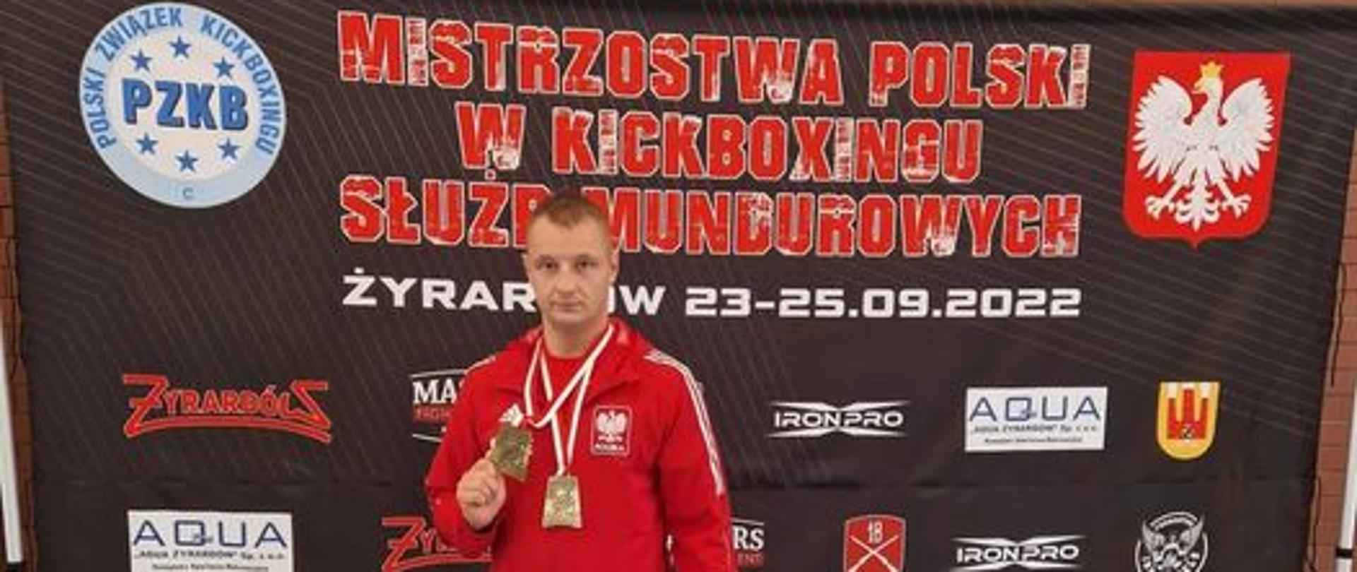 Strażak z medalem w ręku stoi na tle baneru z napisem Mistrzostwa Polski Wojska Polskiego i Służb Mundurowych w kickboxingu