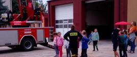 Grupa dzieci wraz ze strażakiem i opiekunem stoją na dworze w pobliżu samochodu strażackiego - drabiny.