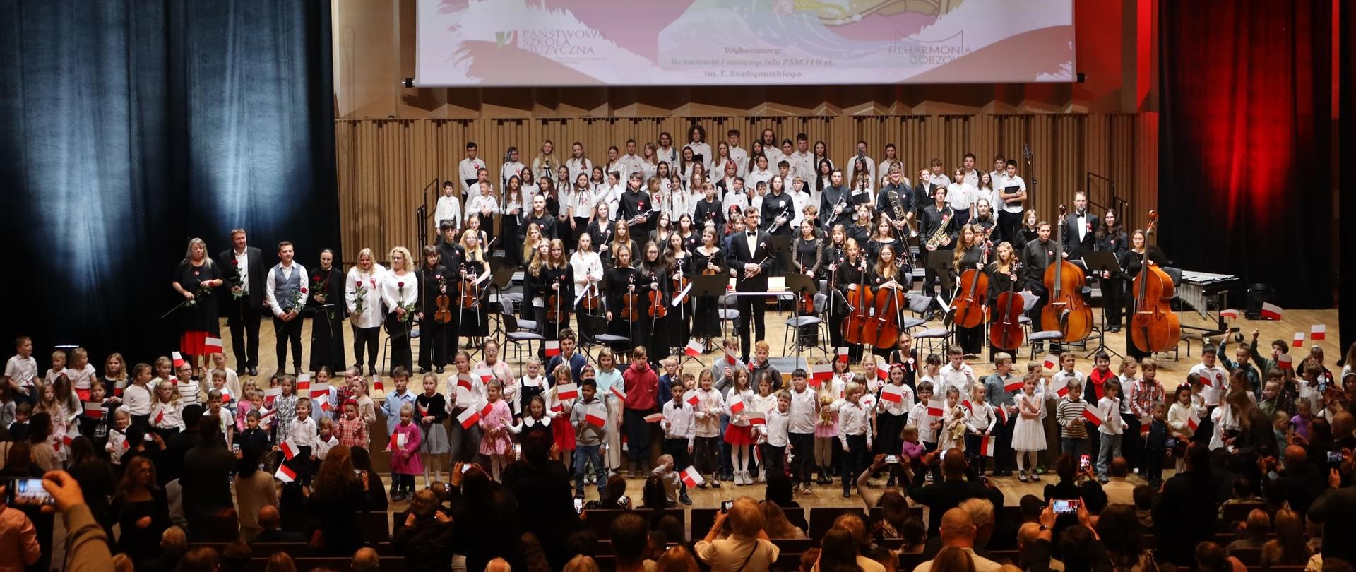 Na scenie filharmonii wykonawcy koncertu - chóry, orkiestry, soliści, dyrygent, prowadzący. Pod sceną dzieci z biało-czerwonymi chorągiewkami