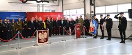 Po lewej stronie stoją za trybuną honorową stoją zaproszeni goście mundurowi ubrani w mundury osoby cywilne w garnitury. W tle czerwony banner Państwowej Straży Pożarnej oraz granatowy baner Związku Ochotniczych Straży Pożarnych. Po prawej stronie poczet sztandarowy.