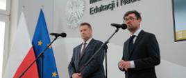 Minister Czarnek i mężczyzna w czarnym garniturze stoją za mikrofonami, za nimi flagi Polski i UE.