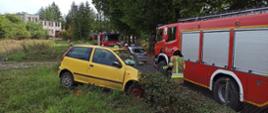 Zdjęcie przedstawia widok żółtego samochodu osobowego, który uderzył w ogrodzenie posesji. Zdjęcie wykonane z dachu samochodu strażackiego. Wokół pojazdu strażacy w ubraniach specjalnych