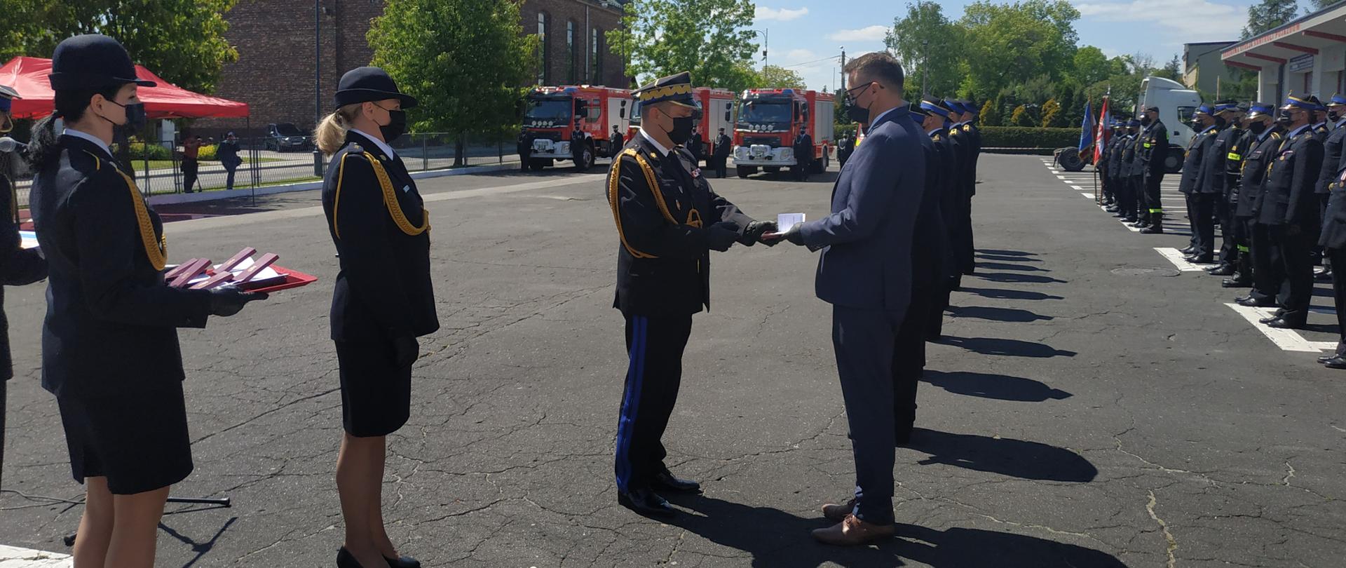 Na tle pododdziałó biorących udział w uroczystej zbiórce z okazji Regionalnych Obchodów Dnia Strażaka 2021 znajdują się osoby odznaczone. Odznaczenia wręczają Śląski Komendant Wojewódzki Państwowej Straży Pożarnej nadbrygadier Jacek Kleszczewski.