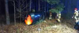 Zdjęcie wykonane w nocy. Na poboczu, poza drogą, leży auto osobowe na boku. W tle las. Po prawej stronie zdjęcia strażacy w piaskowych mundurach.