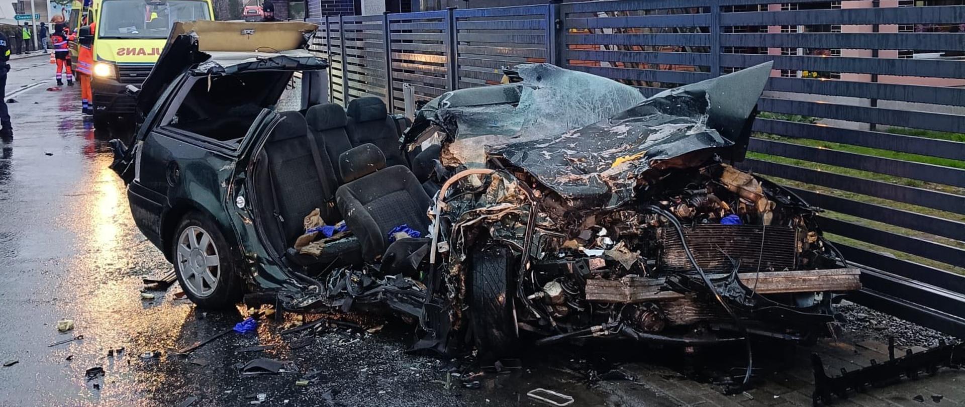 Samochód osobowy po bardzo poważnym wypadku stoi na mokrej jezdni obok metalowego ogrodzenia. Przednia część pojazdu jest całkowicie zniszczona, z odsłoniętymi elementami silnika i pogiętą konstrukcją. Przednia szyba jest rozbita, a dach częściowo usunięty, prawdopodobnie przez służby ratownicze. W tle widoczne są pojazdy ratunkowe, w tym karetka, oraz ratownicy pracujący na miejscu zdarzenia