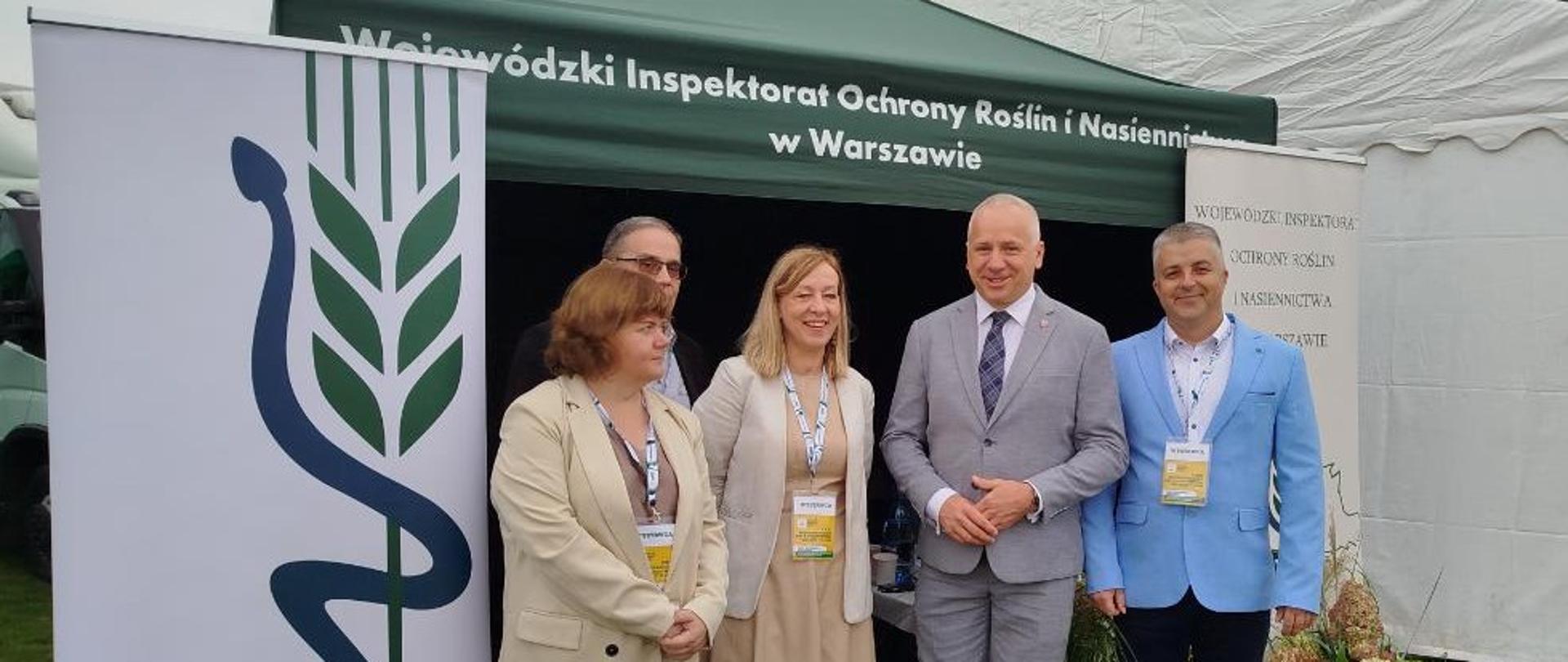 Na zdjęciu widzimy grupę pięciu osób stojących przed namiotem z napisem „Wojewódzki Inspektorat Ochrony Roślin i Nasiennictwa w Warszawie”. Obok widoczne są roll-upy z logo PIORiN oraz dekoracje z kwiatami. Uczestnicy są elegancko ubrani i noszą identyfikatory, pozując do wspólnego zdjęcia.