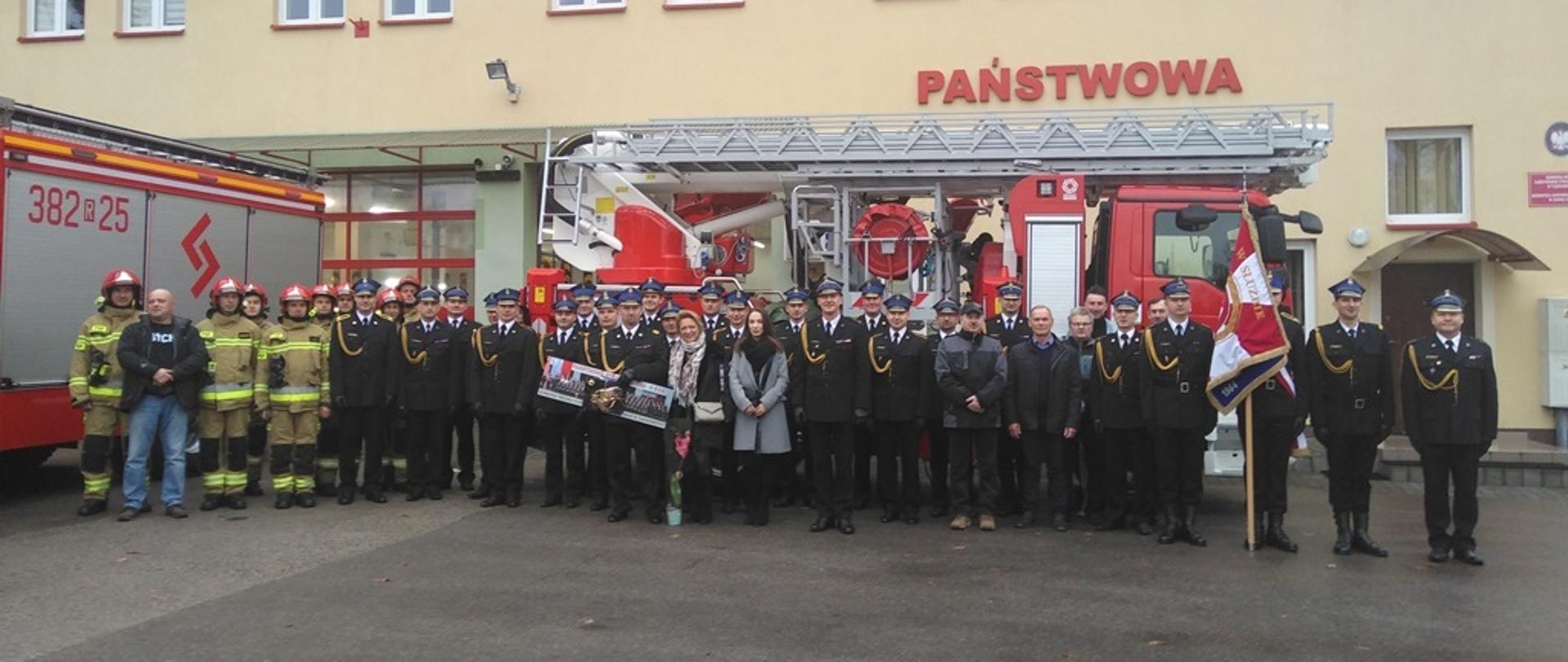 Na placu przed budynkiem remizy Jednostki Ratowniczo-Gaśniczej w Nowej Dębie, przed samochodem z drabiną stoją uczestnicy uroczystej zbiórki. W śród stojących, po prawej stronie fotografii, widoczny sztandar, który prezentuje poczet sztandarowy składający się z trzech strażaków. Po lewej stronie fotografii stoją strażacy w umundurowaniu bojowym w hełmach. W środku widoczny strażak trzymający pamiątkową fotografię, obok którego stoją dwie kobiety oraz strażacy w umundurowaniu wyjściowym. 