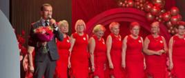 Wicewojewoda w ciemnym garniturze przemawia na scenie. Za nim stoją seniorki w czerwonych sukienkach.