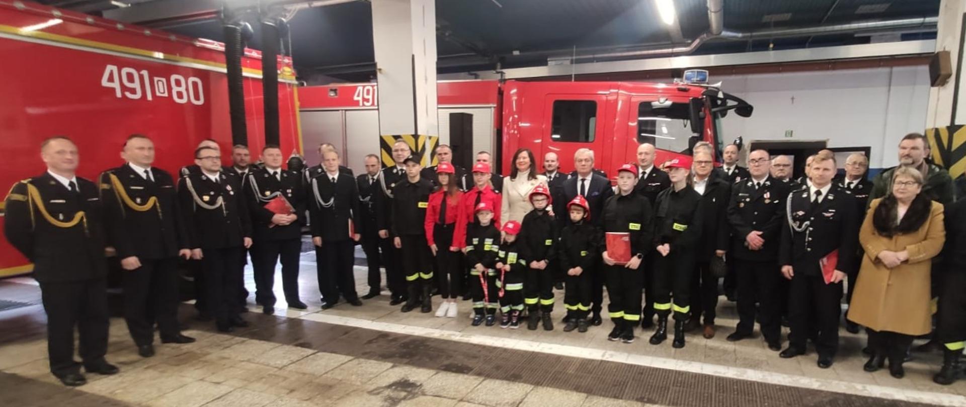 Na zdjęciu znajdują się strażacy oraz osoby zaproszone, ustawione na garażu na tle pojazdów strażackich