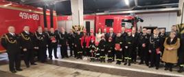 Na zdjęciu znajdują się strażacy oraz osoby zaproszone, ustawione na garażu na tle pojazdów strażackich