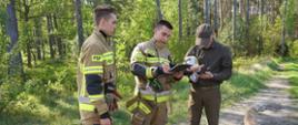 Na drodze leśnej stoi dwóch strażaków w ubraniach specjalnych w kolorze piaskowym bez nakrycia głowy. Jedenz nich trzyma w ręce czerwony hełm. Drugi trzyma wrękach podkładke z dokumentami, w których dokonuje zapisów. Obok stoi leśniczy przeglądający telefon. Na drodze siedzi pies.